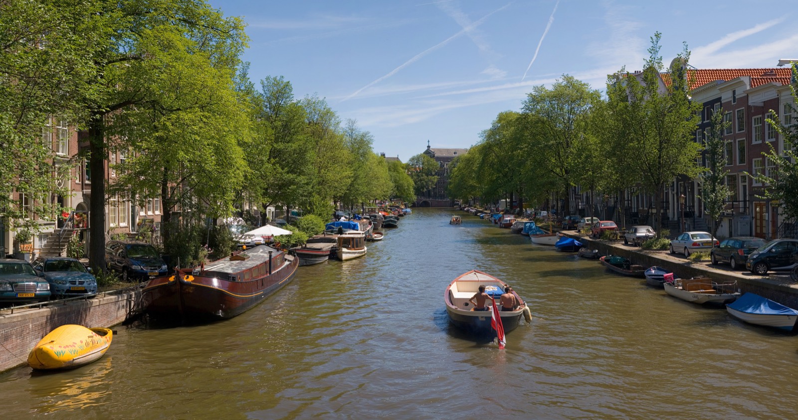 Panorama of an Amsterdam canal in summer, with gabled houses, trees and moored boats.